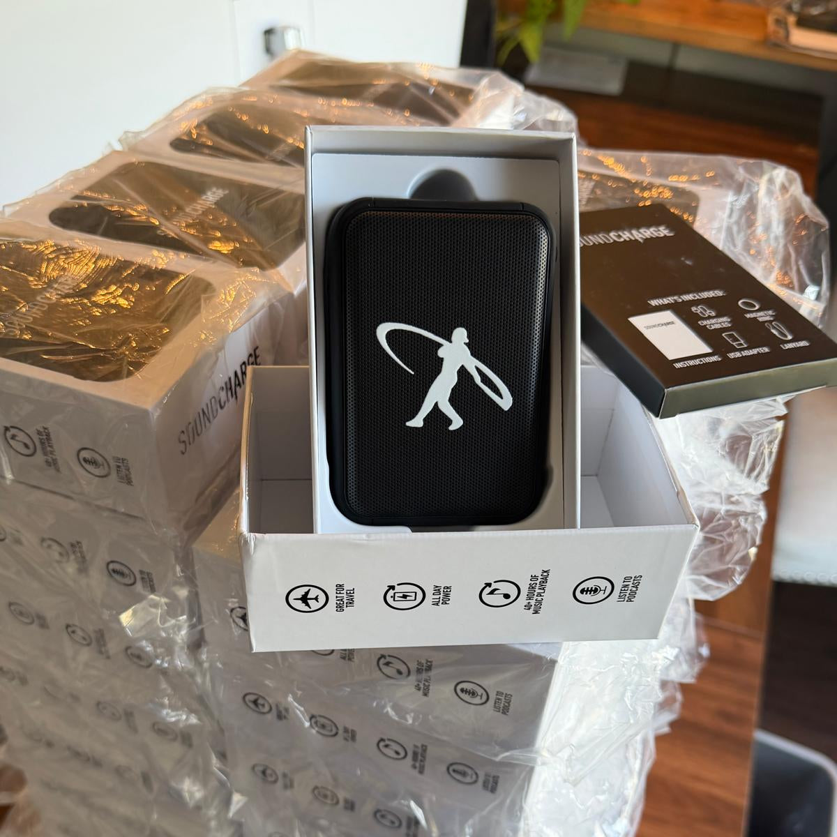A black rectangular electronic device with a white silhouette logo sits in an open white product box, surrounded by stacked shipping boxes wrapped in clear plastic on a wooden table.